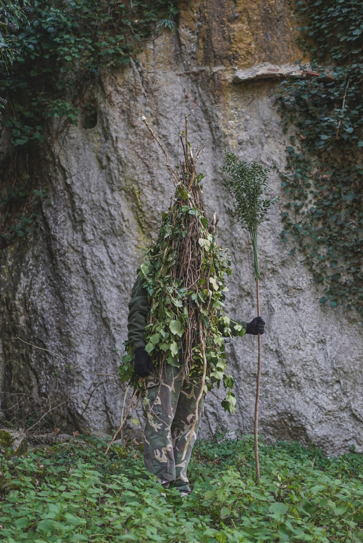 A photograph of a man wearing a “Rumita” costume (made from tree branches) standing in the middle of a forest, holding the typical stick used for the ritual, called a “fruscio”
