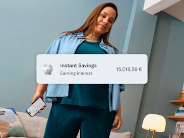 Woman dressed in blue, sitting, and holding her cell phone with the N26 application open. In the foreground, image of the 1.7% p.a. interest rate, balance in the main account, and in the savings account.